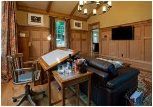 Living room with television on the wall and an drafting table behind a brown sofa,.