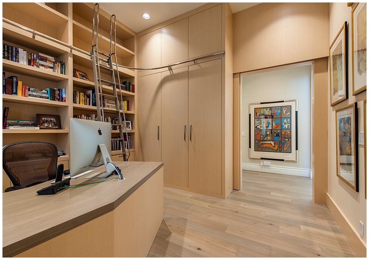 A home office with built-in library wall to the left and a moving ladder. Office has a large door-less doorway that shows a big piece of artwork.