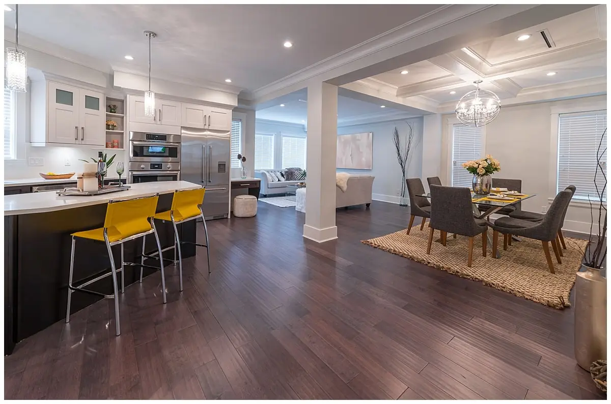 An open place space. Left side has a kitchen breakfast bar with yellow stools. Right side is a formal dining area with dark brown dining chairs.